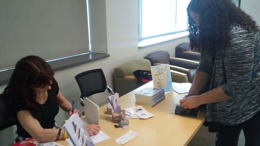 A woman and author signs a book for a reader sitting at a table in a library with pens, a money box, and more books on the table.