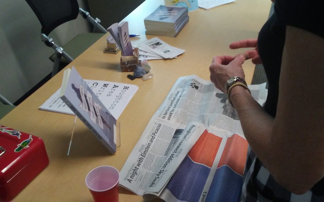 An author's hands holding a pen with a Michigan Tech Lode and a cash box and bookmarks on the table in the library of a University at a book signing.