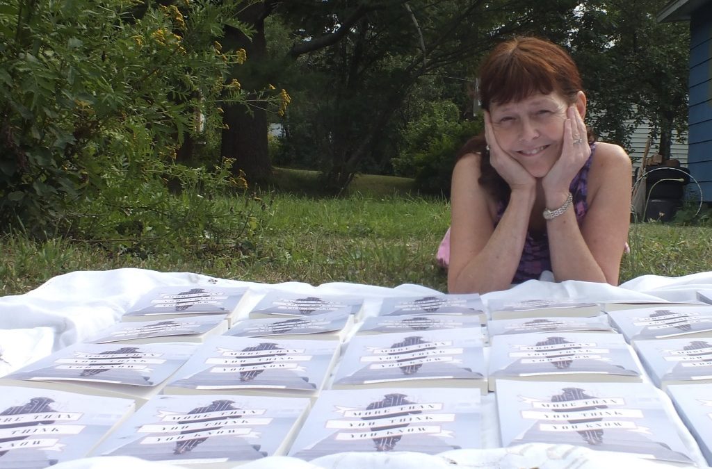 A woman lying on the ground with her chin propped on elbows and hands outside in the summer, with copies of More Than You Think You Know the novel laid out symmetrically before her on the ou