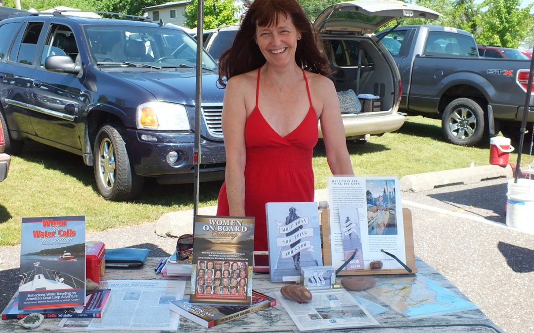 A smiling woman in a read dress at a book festival in Chassell with her novel More Than You Think You Know, Michigan Tech Magazine copies, When the Water Calls We Follow, and Women on Board Cruising