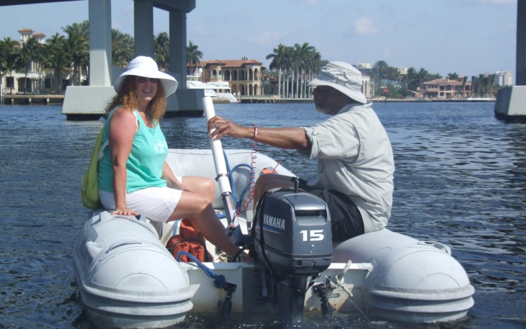 A woman and a man wearing sun hats sitting on the pontoons of their inflatable dinghy going under a bridge with an outboard engine on the dinghy