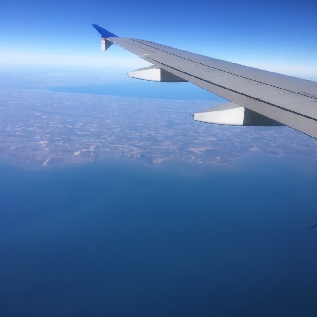 The wing of a plane visible with the blue of a large river below and a land mass in the distance.