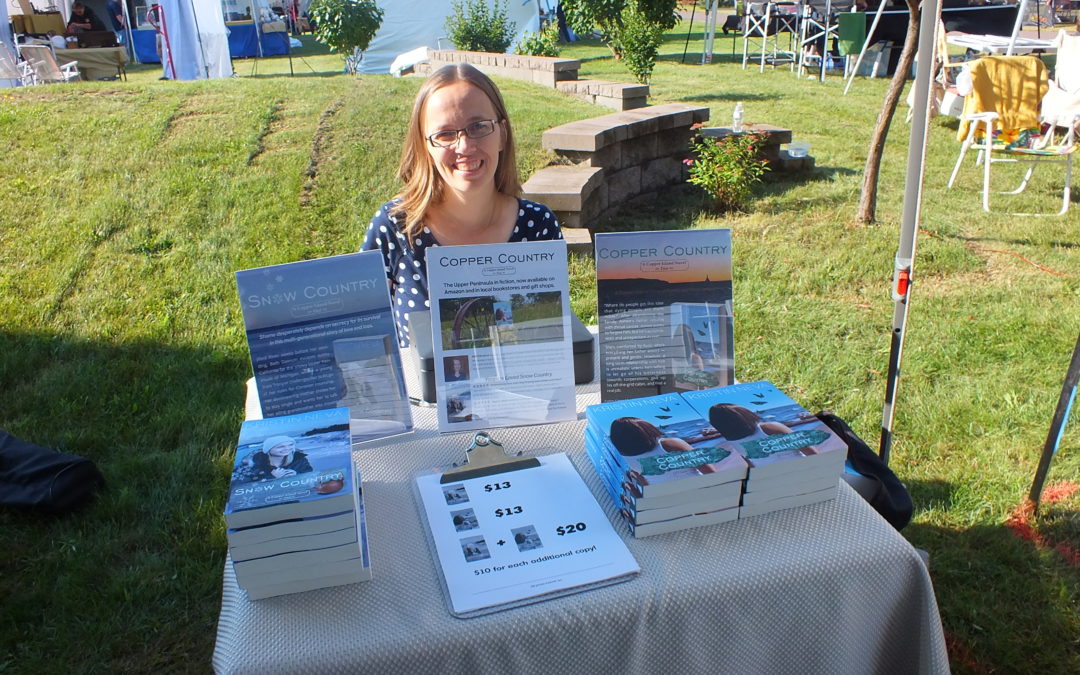 Kristin Neva author with four books sitting at a table at an outdoor art fair under a white tent